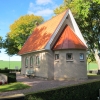 En liten idyllisk byggnad bland högväxta lövträd med välskött kyrkogård.